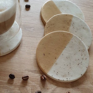 May include: Set of four round coasters with a beige and white speckled design. Each coaster is divided in half, with one half beige and the other half white.