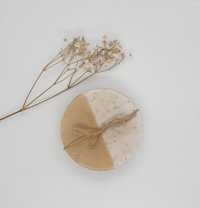 May include: A round ceramic coaster with a beige and white speckled glaze. The coaster is tied with a piece of twine.