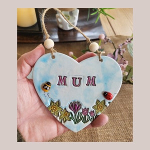 May include: A blue heart-shaped ceramic sign with the word "MUM" in pink letters. The sign is decorated with yellow daffodils, pink tulips, green leaves, a ladybug, and a bee.