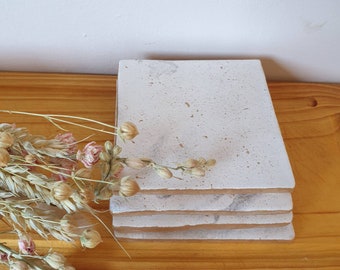 May include: A stack of four white ceramic coasters with a speckled finish. The coasters are square and have a slightly rough texture.