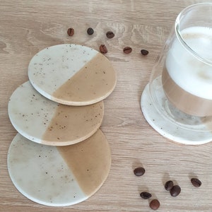 May include: Set of three round coasters with a white and beige speckled design. The coasters are stacked on top of each other. Coffee beans are scattered around the coasters.