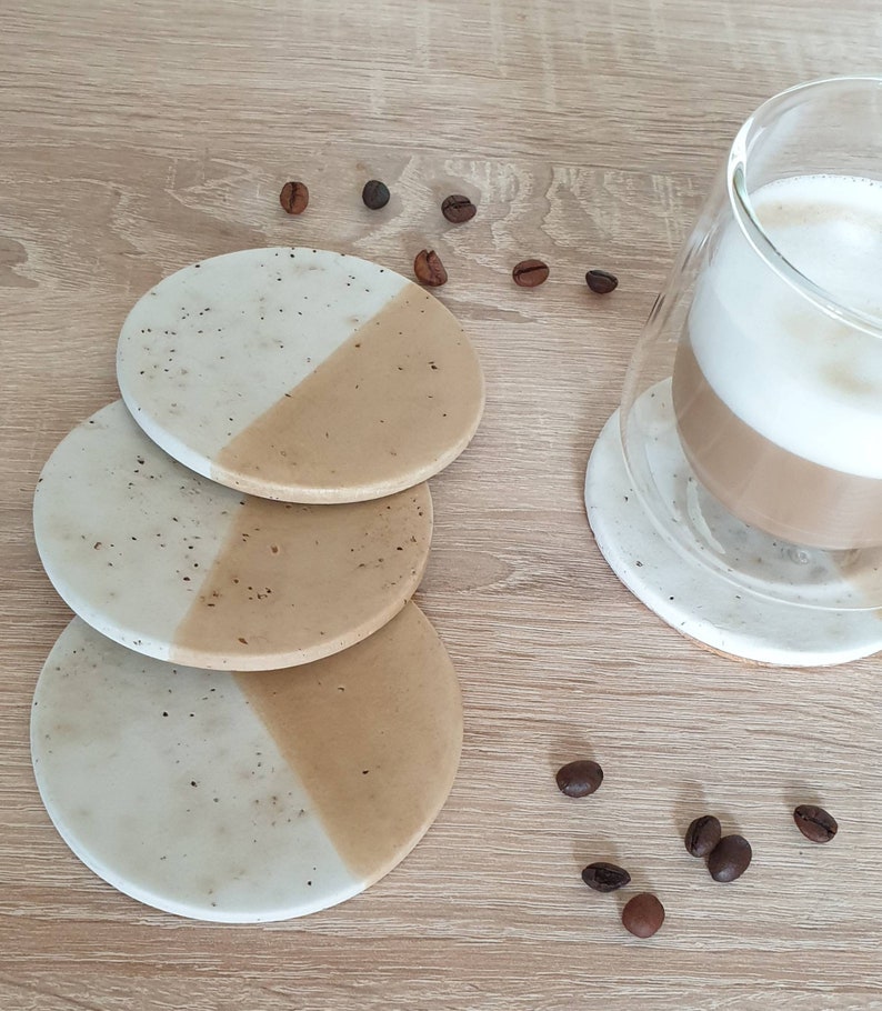 May include: Set of three round coasters with a white and beige speckled design. The coasters are stacked on top of each other. Coffee beans are scattered around the coasters.