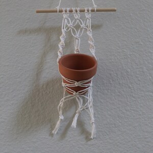 May include: A handmade macrame plant hanger with a small terracotta pot. The off-white macrame is knotted around a wooden dowel and supports the pot. The pot is a warm terracotta color, perfect for small plants.