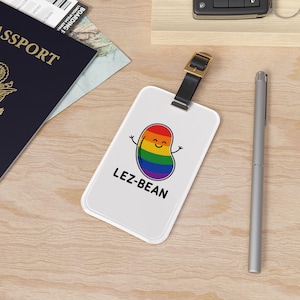 May include: White luggage tag with a black strap and gold buckle. The tag features a cartoon bean design in rainbow colors with the text "LEZ-BEAN". A silver pen and a passport are also visible.