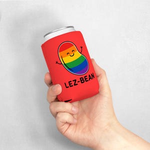 Peut inclure: Un refroidisseur de canette rouge avec le texte "LEZ-BEAN" et un dessin animé de haricot aux couleurs de l'arc-en-ciel. Le haricot a un visage souriant et des bras. Un canette argentée est à l'intérieur.