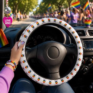 May include: White steering wheel cover with a repeating pattern of rainbow-colored lesbian symbols and the text "LES-BEAN". The image shows a person driving a car, with a "LOVE WINS" heart-shaped charm hanging from the rearview mirror.