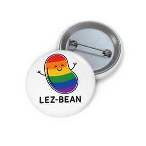 May include: A white button featuring a smiling, rainbow-colored bean character with outstretched arms. The text "LEZ-BEAN" is printed in black below the bean. Another button is visible in the background.