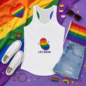 May include: White tank top with a rainbow bean graphic and the text "LEZ-BEAN". The image includes a rainbow flag, denim shorts, white sneakers, and pride-themed accessories.