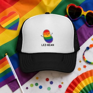 May include: A black and white trucker hat with a white front panel featuring a rainbow-colored bean character and the text "LEZ-BEAN." The hat is set against a rainbow flag background, with rainbow-themed accessories.