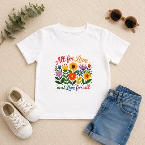 Puede incluir: Camiseta blanca con un diseño floral colorido y el texto "All for Love and Love for all". La imagen incluye pantalones cortos de mezclilla azules, gafas de sol y zapatillas deportivas. También se ve una ramita de vegetación.