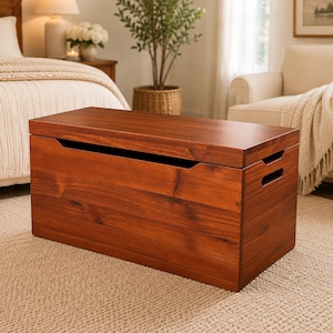 May include: A rectangular wooden storage chest with a warm brown finish and visible wood grain. The chest features a flat top lid, a recessed front opening for easy access, and integrated cutout handles on the side panels for portability.