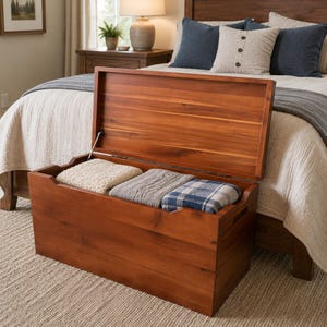 May include: A rectangular wooden storage chest with a hinged lid in an open position. The chest features a warm brown wood grain finish and contains three folded blankets with different textures and patterns, including cream, gray, and blue plaid.