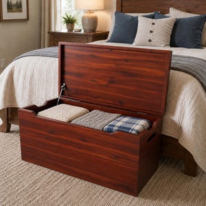 May include: A rectangular wooden storage chest with a rich reddish-brown finish and a hinged lid that is propped open. Inside the chest, three folded blankets are neatly arranged, featuring cream, grey, and blue plaid patterns respectively.