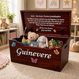 May include: A dark wood toy chest with a hinged lid featuring the name Guinevere in white script and colorful butterfly decals. The interior lid displays a printed message to a granddaughter. The chest contains a teddy bear, a plush rabbit, a wooden house, a stacking ring toy, and a bead maze.