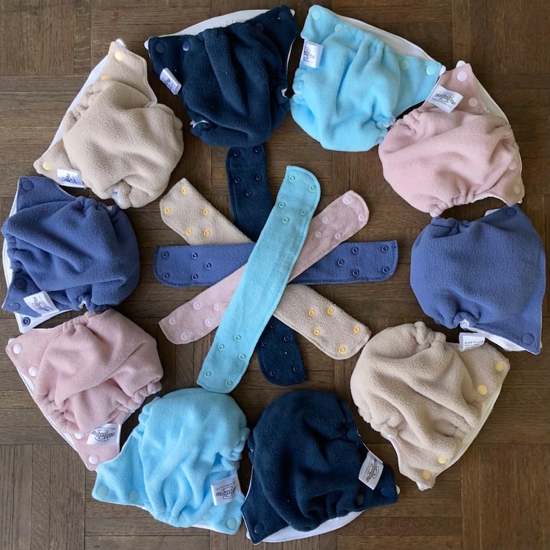 May include: A collection of 10 reusable cloth diapers in various colors, including blue, beige, and light blue. The diapers are arranged in a circle with matching snap closures.