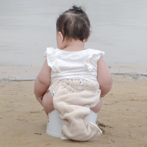 May include: A baby wearing a white shirt and a beige cloth diaper sits on a white potty chair on a sandy beach.