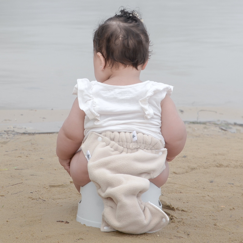 May include: A baby wearing a white shirt and a beige cloth diaper sits on a white potty chair on a sandy beach.