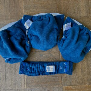 May include: Three blue cloth baby diaper covers with white elastic and blue snaps. The covers are laid out on a wooden surface. The covers have a white label with the text "bumGenius" printed on them.