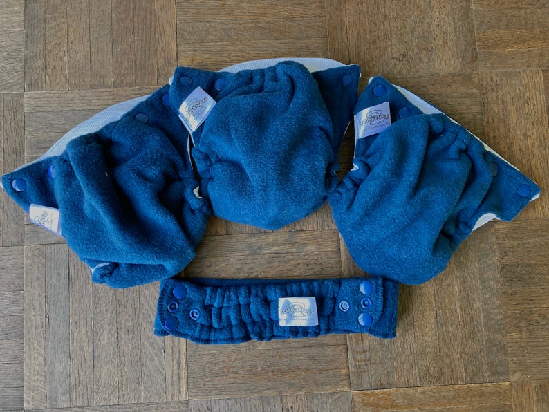 May include: Three blue cloth baby diaper covers with white elastic and blue snaps. The covers are laid out on a wooden surface. The covers have a white label with the text "bumGenius" printed on them.