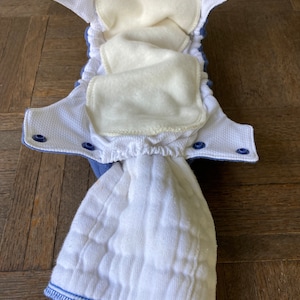 May include: A white cloth diaper with blue trim and snaps. The diaper is open and the inside is lined with a white fleece material.