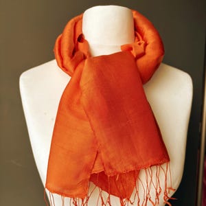 May include: An eye-catching orange silk scarf displayed on a white mannequin. The scarf has a plain design with a slight shimmer, and the edges are trimmed with fine fringe. A fashion accessory.