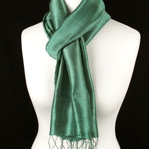 May include: A teal green silk scarf draped over a white mannequin. The scarf has a textured appearance and is knotted at the neck. The ends of the scarf have fringe detailing. The background is black.
