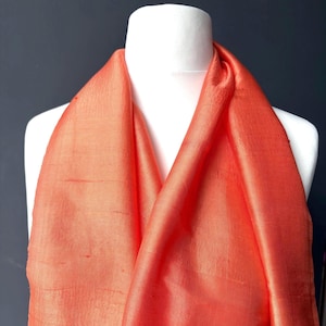 May include: An orange scarf draped over a white mannequin. The fabric has a subtle sheen, catching the light and highlighting its texture. The scarf is made of a lightweight material, ideal for warmer weather.