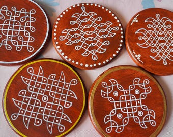 May include: Five circular wooden coasters with a reddish-brown finish, each featuring a unique white hand-painted geometric kolam pattern. The designs include intricate lines, dots, and loops, with some coasters accented by thin yellow or gold painted rims.