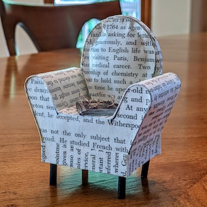 May include: A miniature armchair made from book pages, with a small pair of glasses resting on a tiny book. The chair has black legs and is placed on a wooden surface. The chair is covered in printed text.