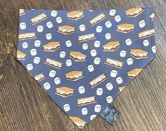 May include: A triangular pet bandana in a dark gray color featuring a repeating pattern of s'mores and white marshmallows. A small black fabric tag attached to the lower right corner displays the white text MagPie Flips in a stylized font.
