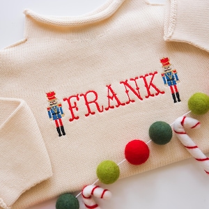 Puede incluir: Un suéter de punto color crema con el nombre "FRANK" bordado en rojo. Dos figuras de soldados de juguete están bordadas en el suéter, una a cada lado del nombre. Una guirnalda de bolas de fieltro rojo y verde y bastones de caramelo está drapeada sobre el suéter.