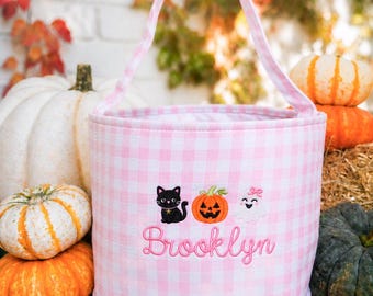 Puede incluir: Una cesta cilíndrica de tela con un patrón de cuadros vichy en rosa claro y blanco y una sola asa. La parte frontal está bordada con un gato negro, una calabaza naranja, un fantasma blanco y el nombre Brooklyn en letra cursiva rosa.