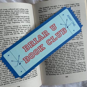 Könnte beinhalten: Ein hellblaues, rechteckiges Lesezeichen in Ticketform mit gezackten Rändern, das den roten Schriftzug BRIAR U BOOK CLUB, zwei gekreuzte Eishockeyschläger in Dunkelblau und den Text ADMIT ONE zeigt, platziert auf einem aufgeschlagenen Buch.