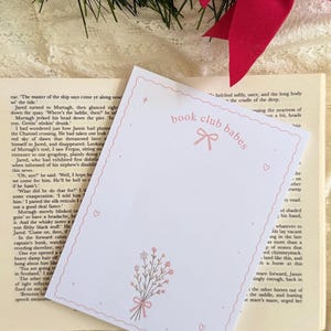 以下が含まれることがあります： ピンクのスカラップ縁取りが施された白い長方形のカード。ピンク色の「book club babes」という文字、ピンクのリボンのグラフィック、リボンで結ばれたピンクの花束が描かれており、開いた本のページの上に置かれています。