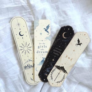 May include: Four rectangular bookmarks featuring celestial and fantasy illustrations. Designs include a crescent moon, stars, a dragon, a sword, and mountains. Text reads "for the girls who dream" and "currently ignoring reality" in black font.
