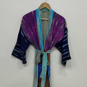 May include: A long-sleeved robe featuring a tie-dye pattern in shades of purple, dark blue, light blue, and rust orange. The garment includes a matching fabric belt at the waist, an open front design, and two front pockets, all hanging on a wooden hanger.
