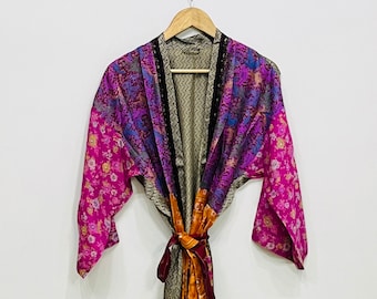 May include: A colorful, patterned kimono robe with a floral design in shades of pink, purple, and orange. The robe is hanging on a wooden hanger against a white background. The robe is tied at the waist with a matching sash.