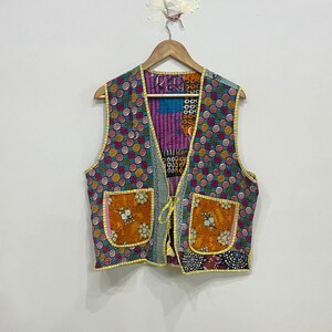 Könnte beinhalten: Eine farbenfrohe, gesteppte Weste mit einem einzigartigen Patchwork-Design. Die Weste hat einen tealfarbenen Hintergrund mit rosa und gelben Kreisen, orangefarbenen Taschen und gelben Zierleisten. Sie hängt an einem Holzbügel vor einer weißen Wand.