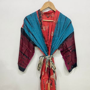 Peut inclure: Robe colorée à motifs, avec fermeture à nouer, suspendue à un cintre en bois. La robe présente un mélange de couleurs, dont le turquoise, le rouge et le bordeaux. Le tissu a un motif de feuilles et une ceinture à nouer.