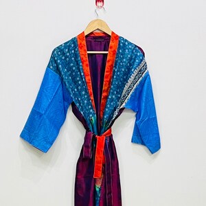 Könnte beinhalten: Ein farbenfroher Morgenmantel in Blau-, Türkis-, Orange- und Lilatönen. Der Morgenmantel hat ein gemustertes Design am oberen Teil und an den Ärmeln, mit einem kontrastierenden orangefarbenen Besatz und einem passenden Gürtel. Er hängt an einem Holzbügel.