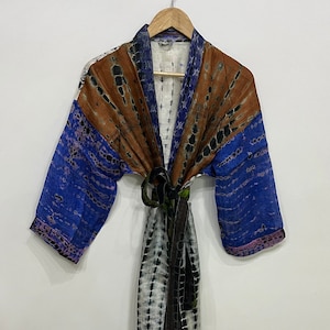 May include: A tie-dye patterned robe in shades of brown, blue, and white hangs on a wooden hanger. The robe features a belt and a long, flowing design. The sleeves are blue with a tie-dye pattern.