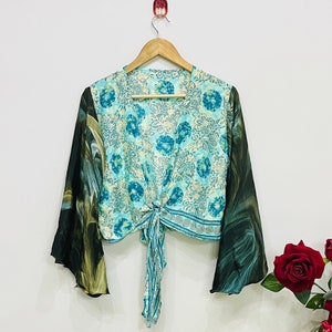May include: A cropped tie-front top featuring a light blue and teal floral pattern on the bodice and long, flowing sleeves with a dark green and black marbled print. The garment is displayed on a wooden hanger against a plain white wall.