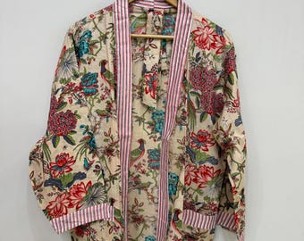 May include: A beige floral print robe with red, blue, and green accents. The robe features a striped pink trim along the edges and sleeves. The robe is hanging on a wooden hanger.