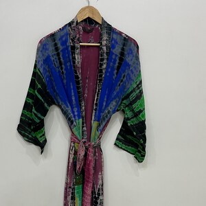 May include: A long-sleeved robe featuring a tie-dye pattern in shades of purple, blue, green, and black. The garment has an open front design with a matching fabric belt at the waist, wide sleeves, and a shawl collar, all hanging on a wooden hanger.