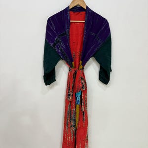 Op de afbeelding: Een kamerjas met lange mouwen en een felrode basis met een tie-dye patroon in zwart, geel en blauw. Het bovenste gedeelte en de mouwen zijn voorzien van donkerpaarse en donkergroene panelen, inclusief een bijpassende ceintuur om te strikken.