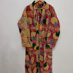 May include: A long-sleeved, open-front robe featuring a vibrant patchwork design with a dominant magenta background. The garment is decorated with repeating circular and triangular patterns in yellow, dark green, and grey, finished with visible stitching.