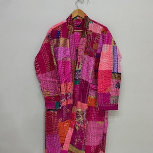 May include: A patchwork robe in shades of pink, magenta, and orange hangs from a wooden hanger. The robe is made of various fabric squares stitched together, creating a unique, textured design. The robe has long sleeves and a wrap-around style.