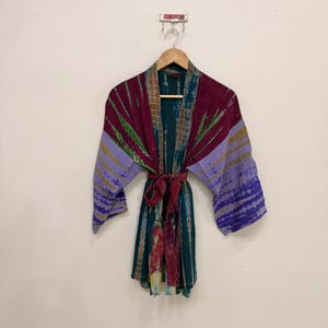 May include: A tie-dye kimono robe featuring a deep burgundy, teal, and lavender color palette. The garment has wide sleeves, an open front design, and a matching fabric tie belt. The fabric displays a horizontal striped pattern with blended colors.