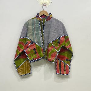 May include: A colorful patchwork jacket with a high collar and long sleeves. The jacket features a mix of patterned fabrics in shades of green, red, and gray, with a blue and white trim. It hangs on a wooden hanger.