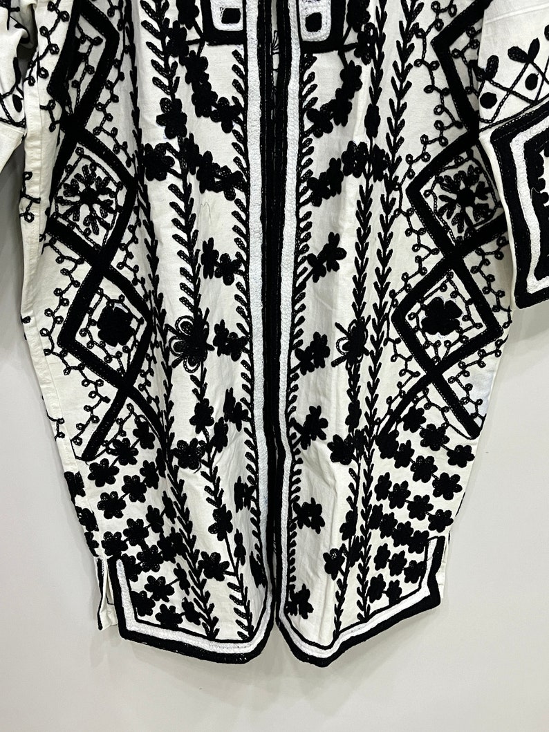 May include: A white cotton tunic with black embroidery featuring a geometric pattern and floral designs.